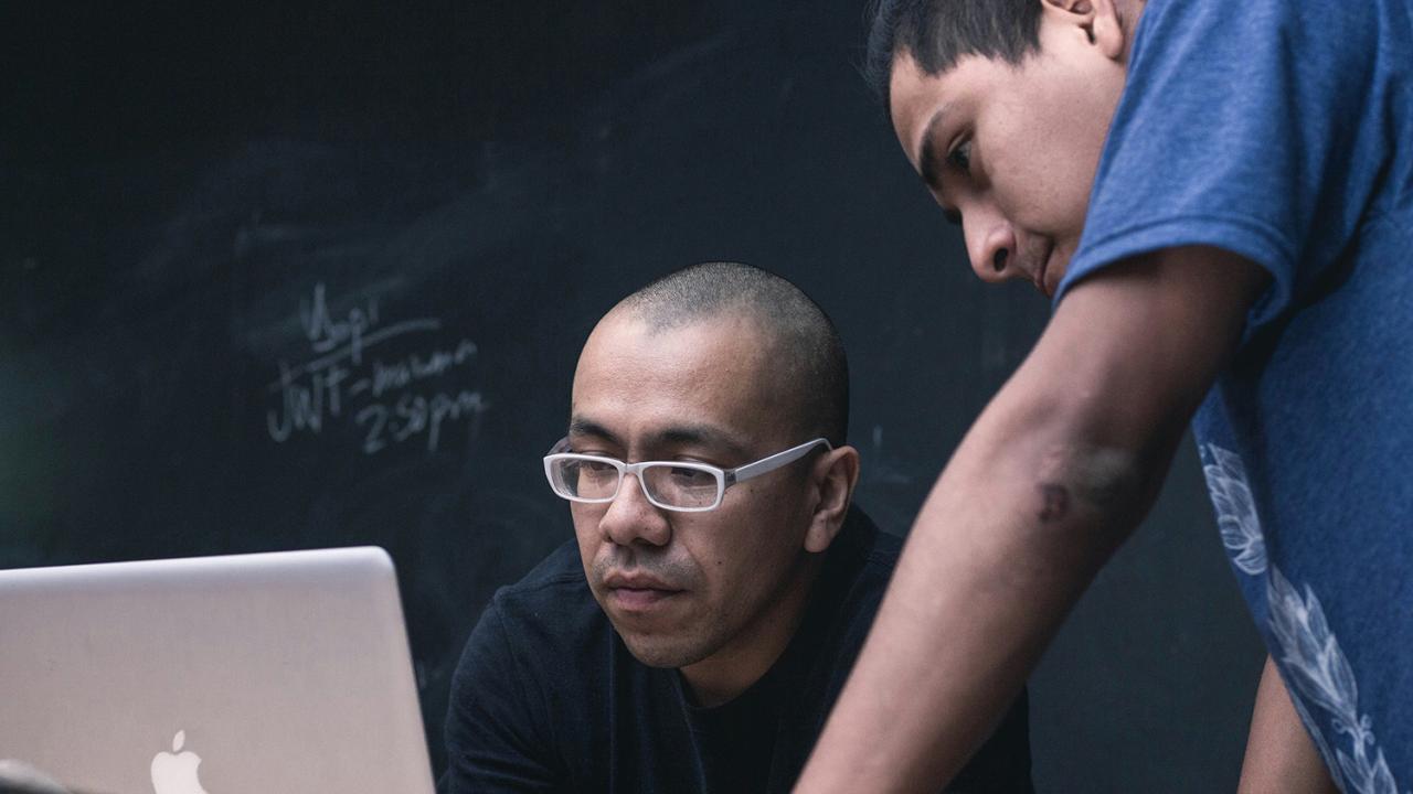 Two people working on a laptop in a classroom.