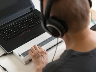 A person wearing headphones uses a laptop with an accessible Braille display.