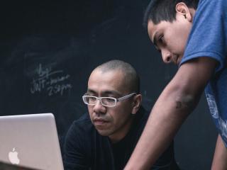 Two people working on a laptop in a classroom.